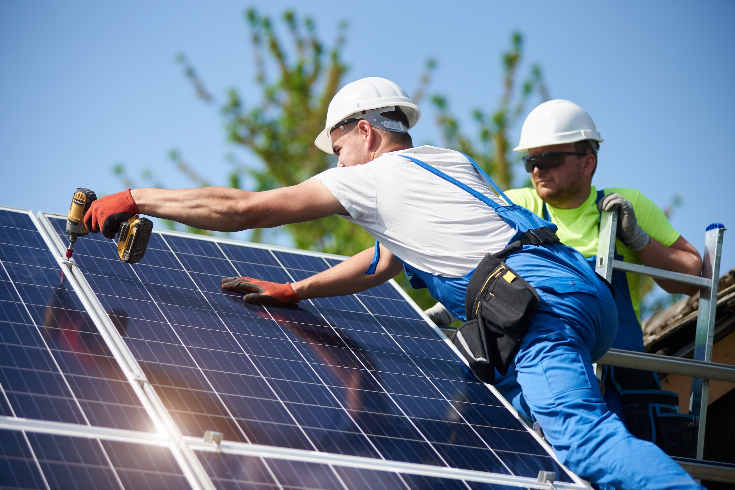 Two solar panel technicians on a roof install a solar panel using a drill.